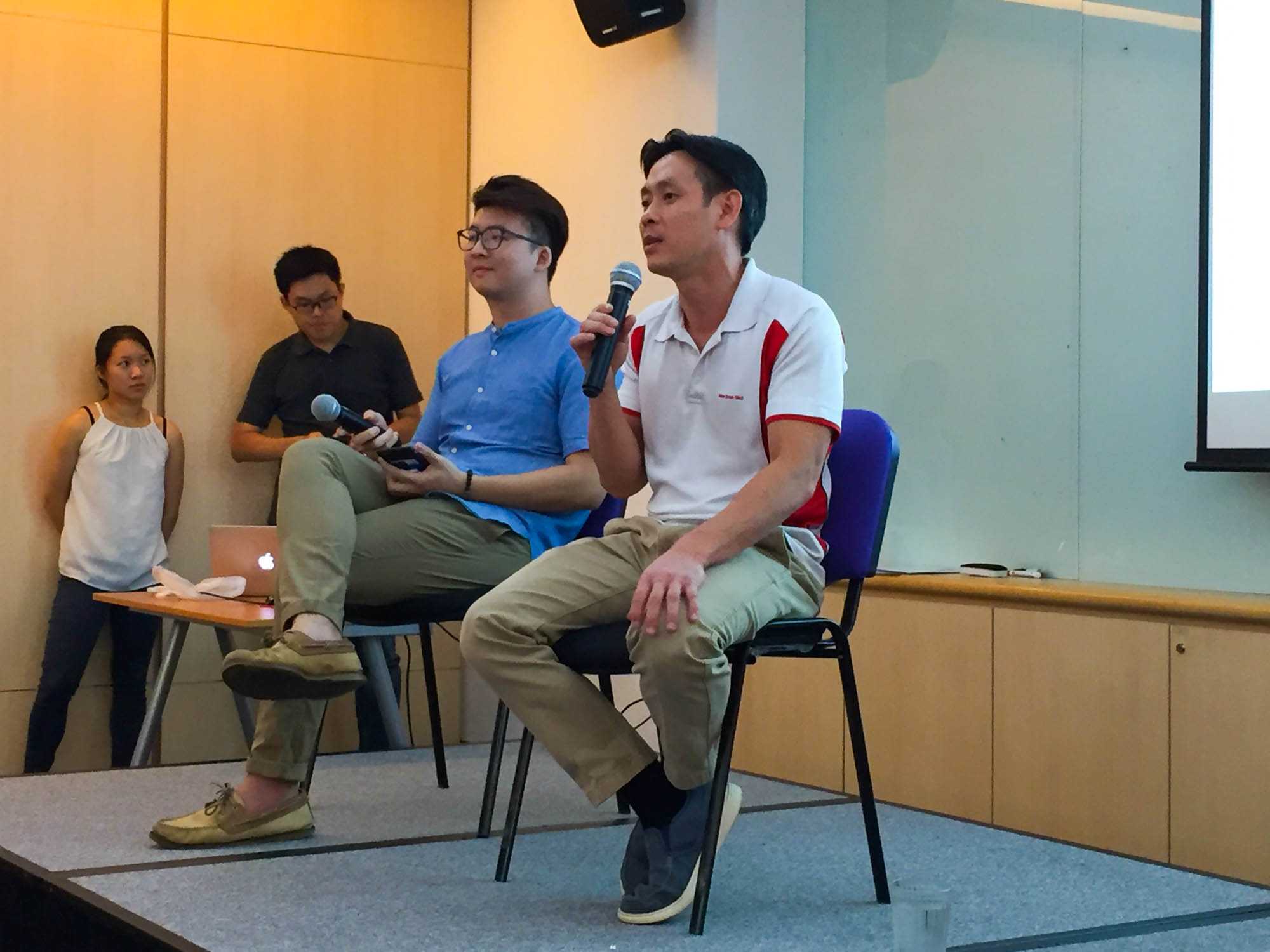 Photo of MP Louis Ng speaking at Budget 101 event, as Rachel stands by the projector handling the slides and audio-visual equipment