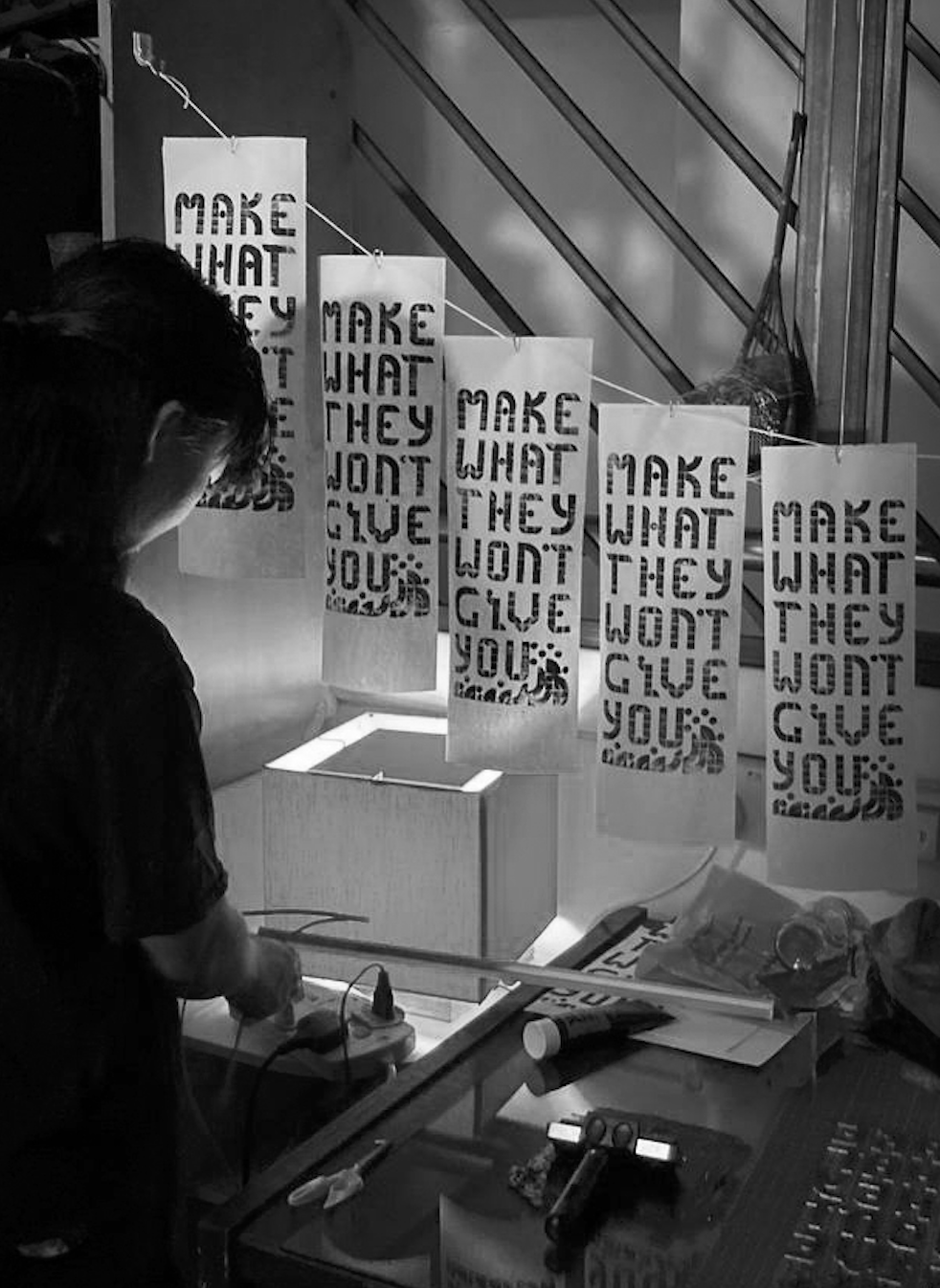 photo of Rachel in her home studio printing a series of posters with the words 'MAKE WHAT THEY WON'T GIVE YOU', using a printing press she made from Legos